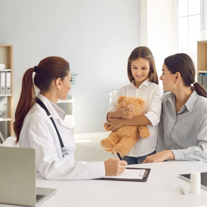 Eine junge Ärztin in ihrem Büro spricht mit einer Frau und ihrer Tochter, die einen Teddybären in den Armen hält.