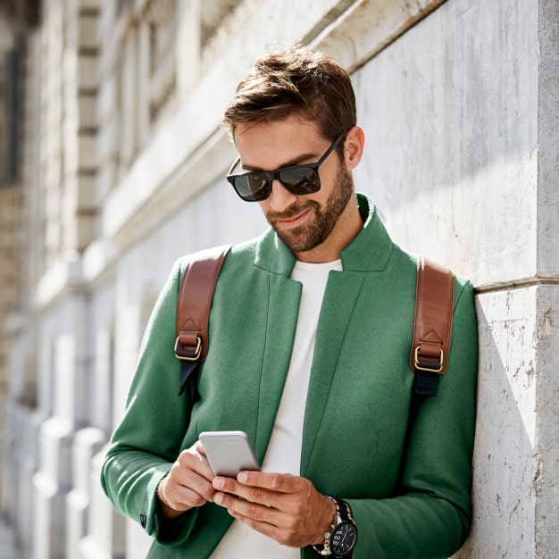 A bearded man in a green blazer with sunglasses is leaning against a wall texting a message in his smartphone