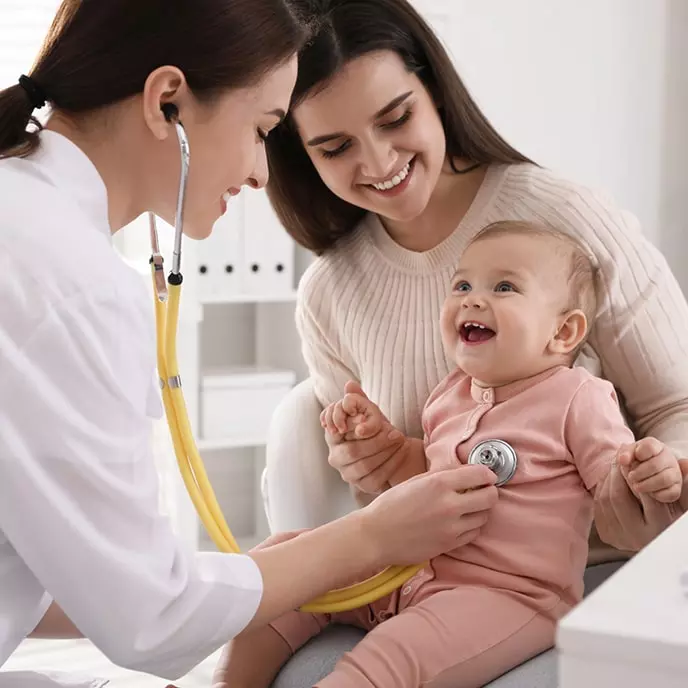 Pediatrs, kurš izmanto stetoskopu, lai izmeklētu smaidīgu mazuli, kas sēž uz mātes ceļiem.